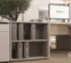 Žema atvira biuro lentyna smėlio spalvos, su dviem lentynomis segtuvams laikyti, šalia darbo stalo su kompiuteriu ir alyvmedžio vazonu.