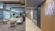 Atviras modernus biuro vestibiulis su Revolut neoniniu “R” ženklu, pilkos sekcijinės sofos ir rausvo atspalvio kėdžių poilsio zona, medžio dekoruotos sienos, stiklinės susitikimų patalpos ir ilgas koridorius su cik-cak grindimis.