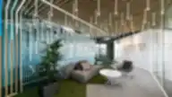 „Helis“ biuro poilsio zona su sofa, moderniu apšvietimu, stiklo pertvaromis ir dirbtinės žolės grindų akcentais.