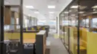 Modernus DHL Lietuva atviro plano biuras su eilėmis darbo stalų, geltonomis akustinėmis pertvaromis ir stiklinėmis kabinomis, kylančiomis iš pilkos ir geltonos kiliminės dangos, tarpai atskirti juodos metalinės grotelės panelėmis.