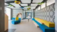 Balsių mokyklos moderni poilsio zona su spalvingais baldais, geometriniais lubų elementais ir dekoruota siena.