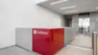 Modernus Hollister biuro interjeras su pilkos spalvos sienomis, ryškiai raudonu prekės ženklo logotipu ir stiklinėmis lifto durimis fone.