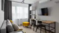 Modernus studijos tipo interjeras Balcia Residence apartamentuose su pilkai geltonais akcentais, valgomojo zona ir kabinamu televizoriumi.
