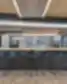 Tiesus „Revolut“ bendros virtuvės baro vaizdas: ilga šviesaus medžio stalviršio sala su matinėmis juodomis subway plytelėmis priekinėje dalyje ir aštuoniais juodais baro taburetėmis; fone – pilkos spintelės su atviromis lentynomis, kuriose išdėstyti balti puodeliai ir stiklinės, įmontuota kavos aparatas ir mikrobangų krosnelė, o dešinėje matyti ryškus samanų akcentas po pakabintomis perforuotomis metalinėmis lubų plokštėmis su LED apšvietimu.