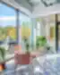 Šviesi SHED Co-living Vilnius poilsio erdvė su vazoniniais augalais ir pastelinėmis kėdėmis, dideliais langais ir išėjimu į terasą su vaizdu į mišką.